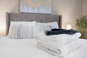 Ein ordentlich bezogenes Bett mit einem gefalteten weißen Handtuch und einem dunkelblauen Handtuch darauf, ein grauer gepolsterter Kopfteil und ein weißes Kissen mit geometrischem Muster.