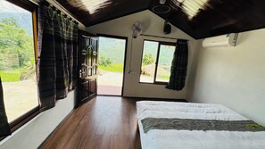 Una habitació amb un llit, una finestra amb cortines, una porta i un ventilador.