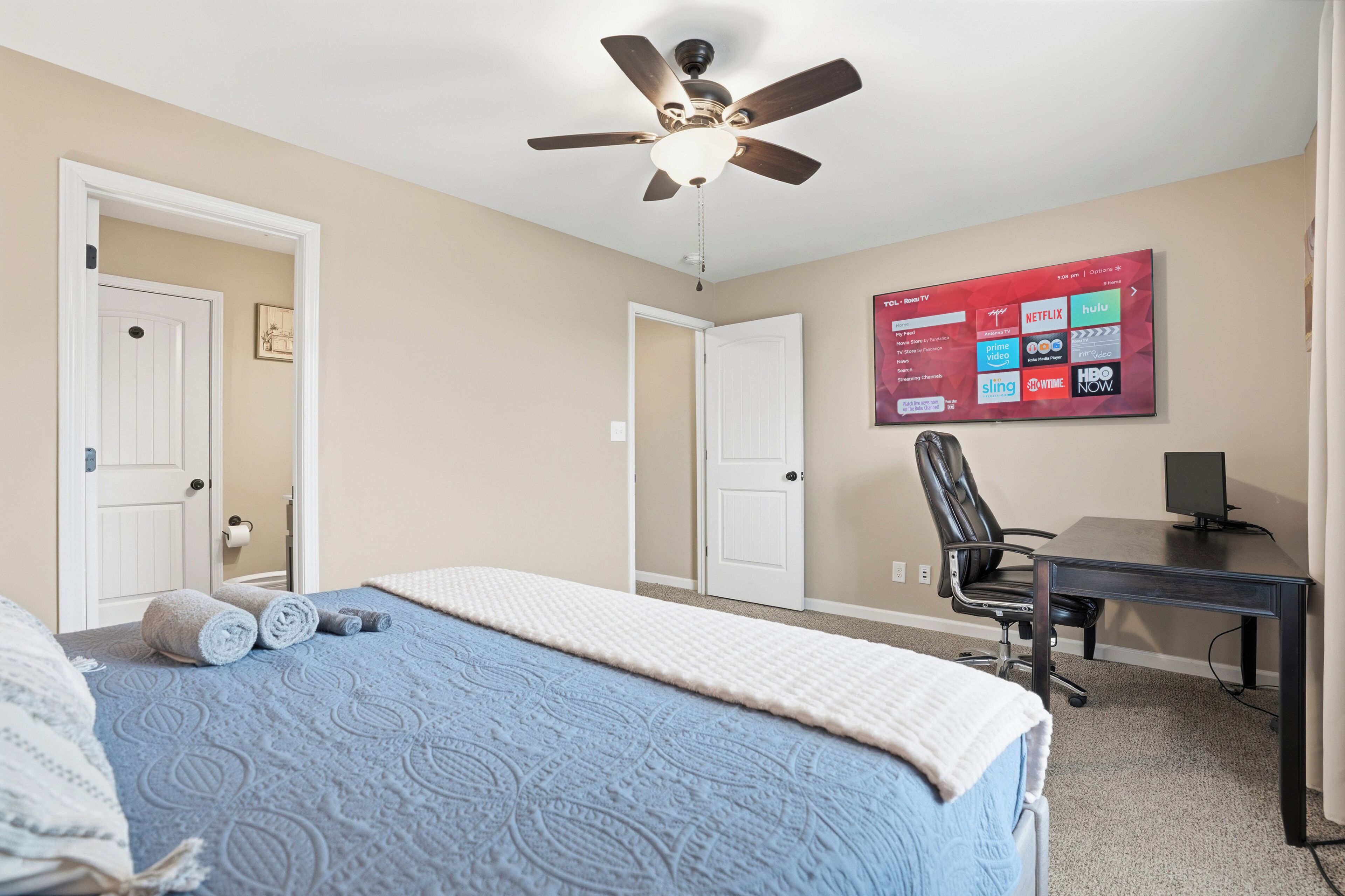 A bedroom with a bed, a ceiling fan, a desk, and a TV displaying streaming services.