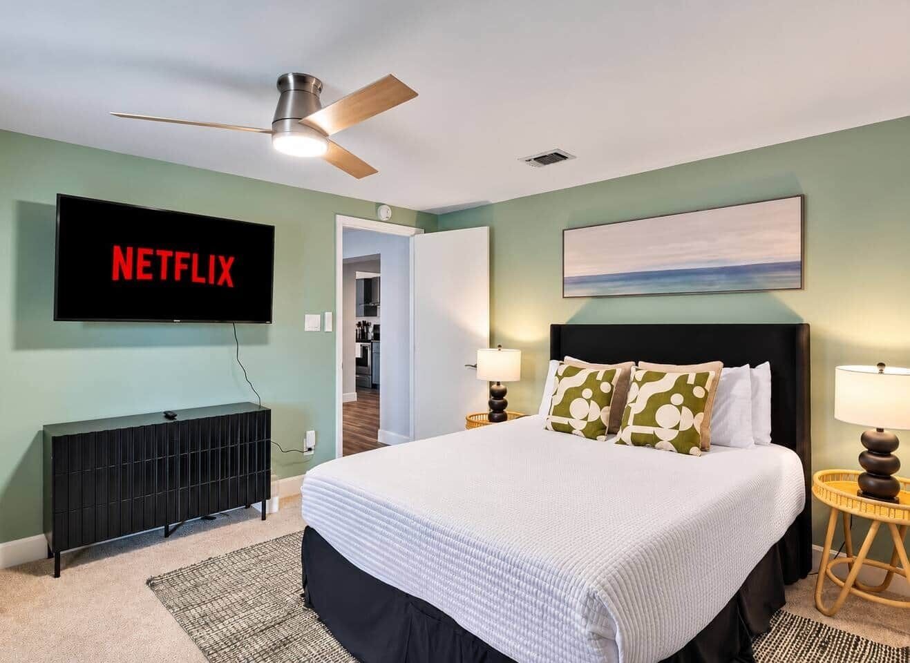 A bedroom with a bed, a TV mounted on the wall, a ceiling fan, and a painting above the bed.