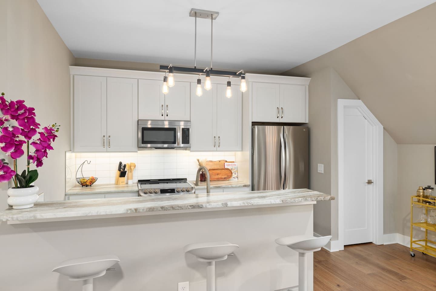 A modern kitchen with white cabinets, a stainless steel refrigerator, and a bar area with stools.