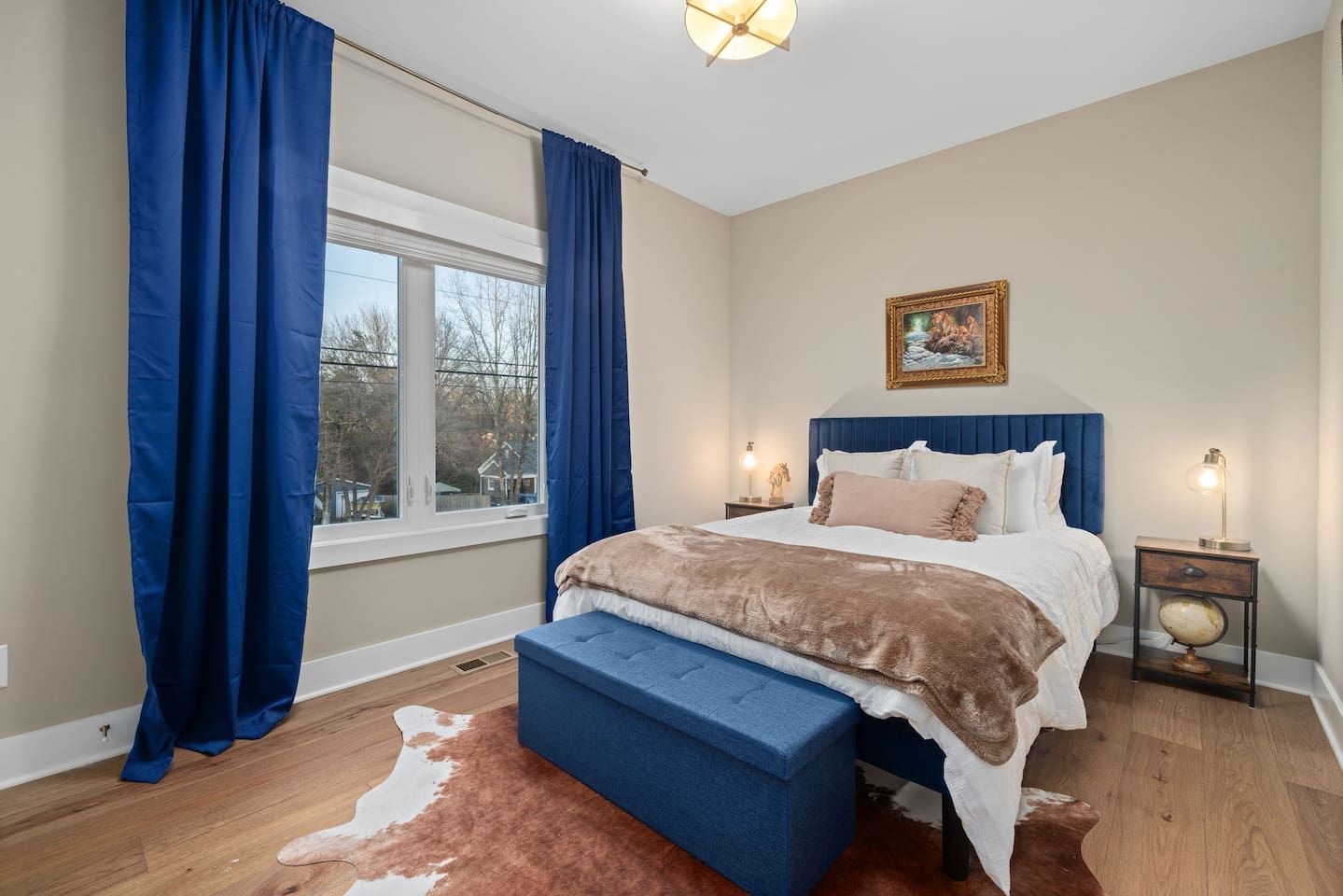 A bedroom with a large bed, a nightstand with a lamp, a window with curtains, and a painting on the wall.