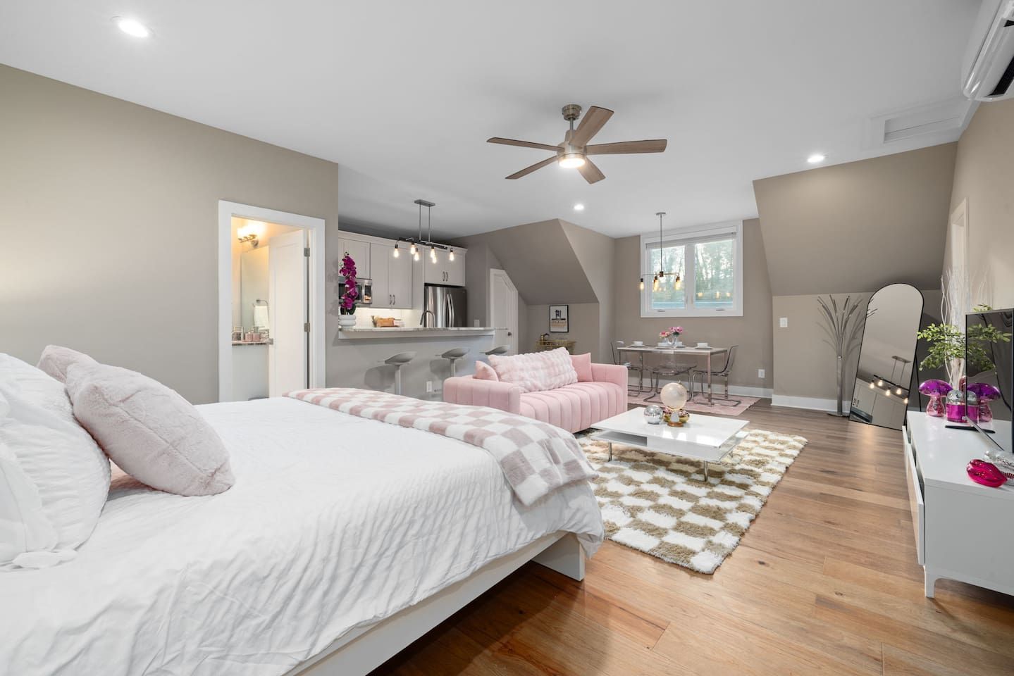 A modern bedroom with a large bed, a pink sofa, a dining area, and a kitchen in the background.