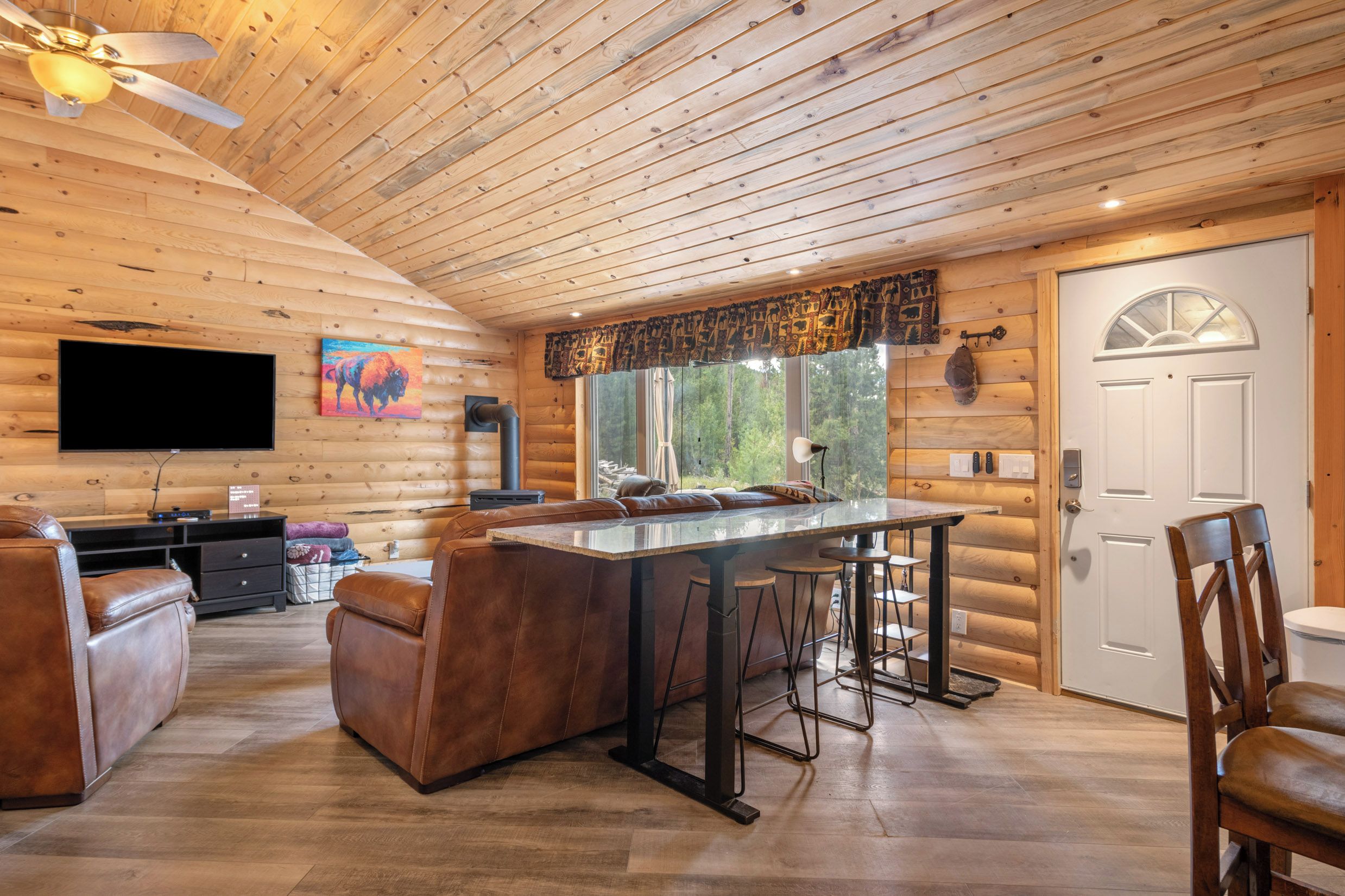 L’intérieur d’une cabane en rondins comprend un coin repas, des sièges en cuir, un téléviseur à écran plat et une grande fenêtre avec des rideaux.