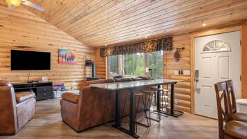 L’intérieur d’une cabane en rondins comprend un coin repas, des sièges en cuir, un téléviseur à écran plat et une grande fenêtre avec des rideaux.