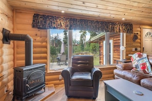 A log cabin interior with a wood-burning stove, leather armchair, and a view of the outdoors.