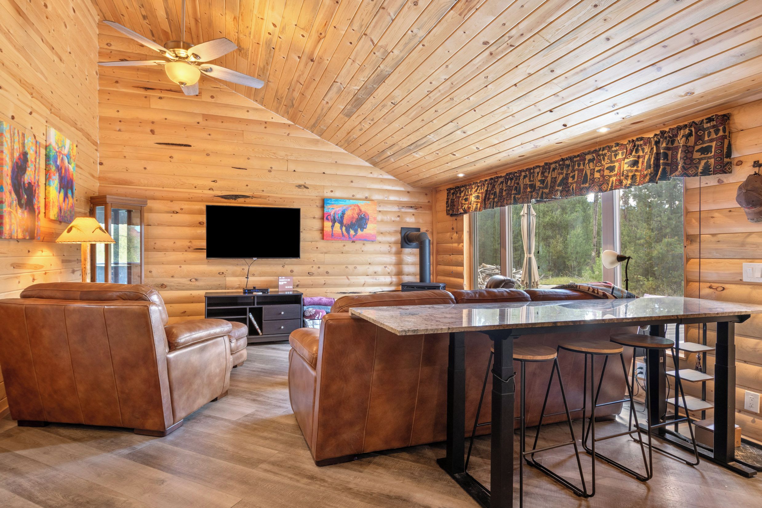 A log cabin interior with a dining area, leather seating, a ceiling fan, and a large window with a view.