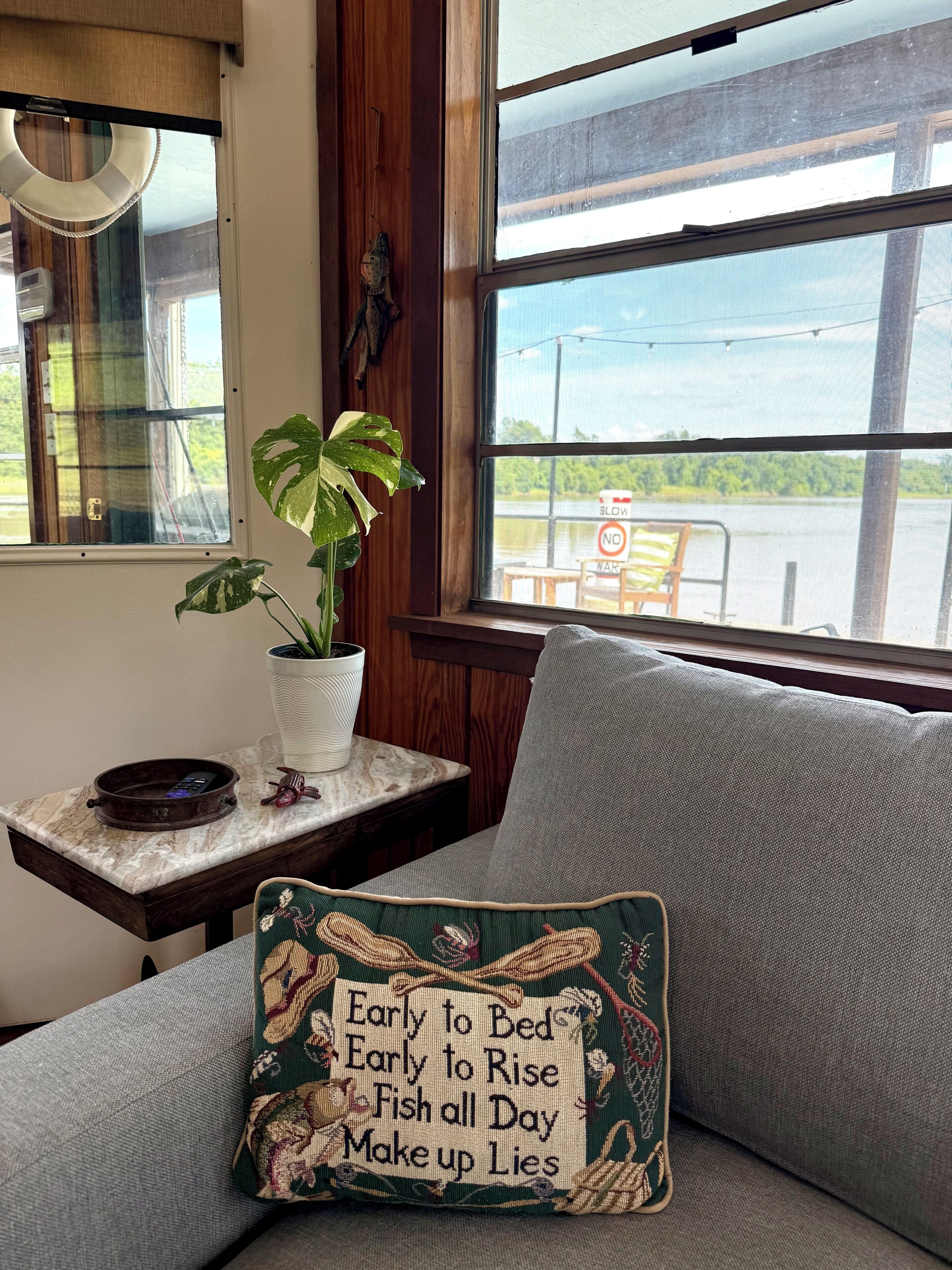 Une pièce avec vue sur un plan d’eau, une plante sur une table et un coussin orné d’une citation amusante.