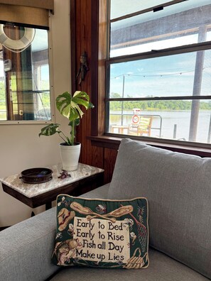 Habitación con vista a un cuerpo de agua, una planta sobre una mesa y un cojín con una cita divertida.