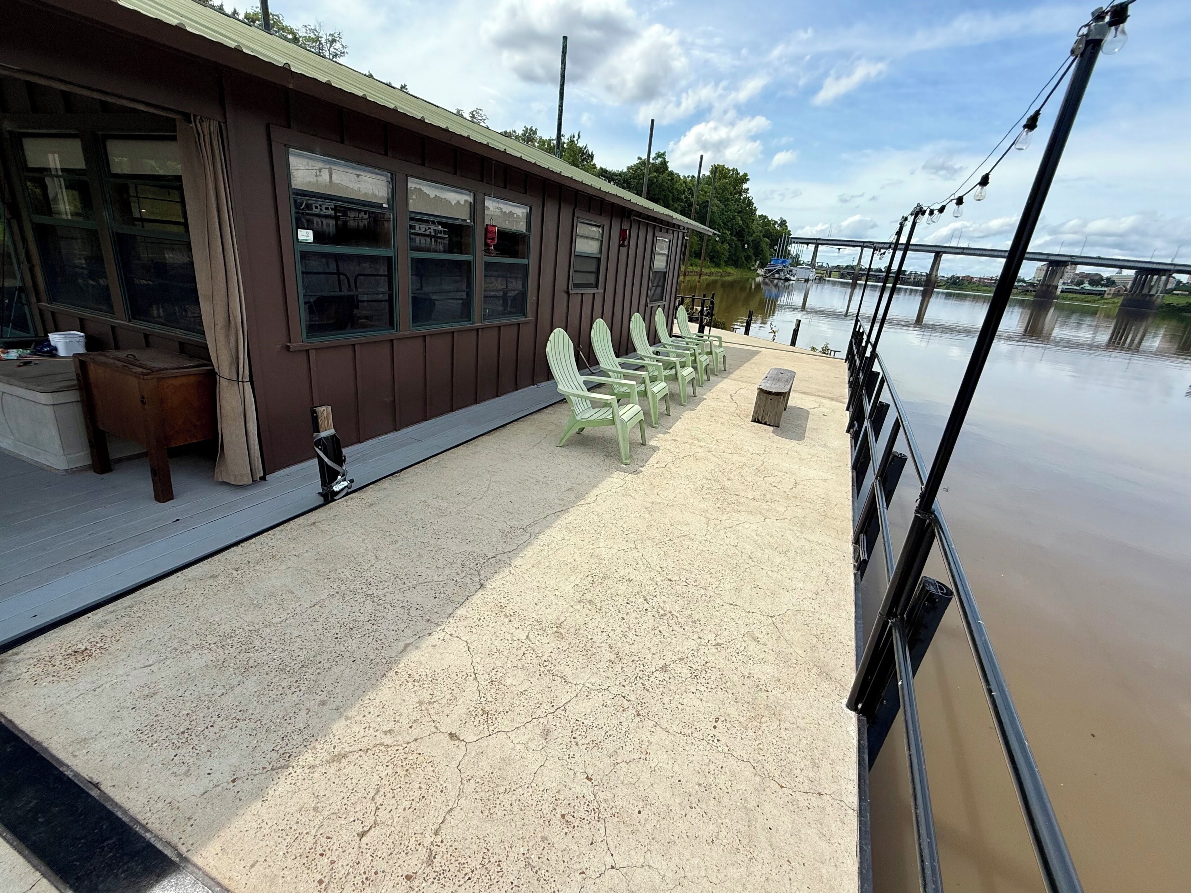 Un bâtiment situé au bord de l’eau, avec une rangée de chaises vertes et un banc, une terrasse en béton et une vue sur une rivière paisible, avec un pont visible au loin.