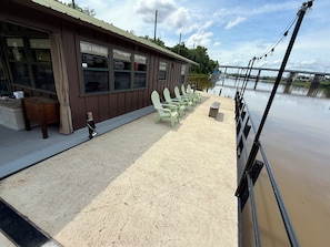 Un edificio frente al río con una fila de sillas verdes y un banco, un muelle de hormigón y vistas a un río tranquilo con un puente a lo lejos.