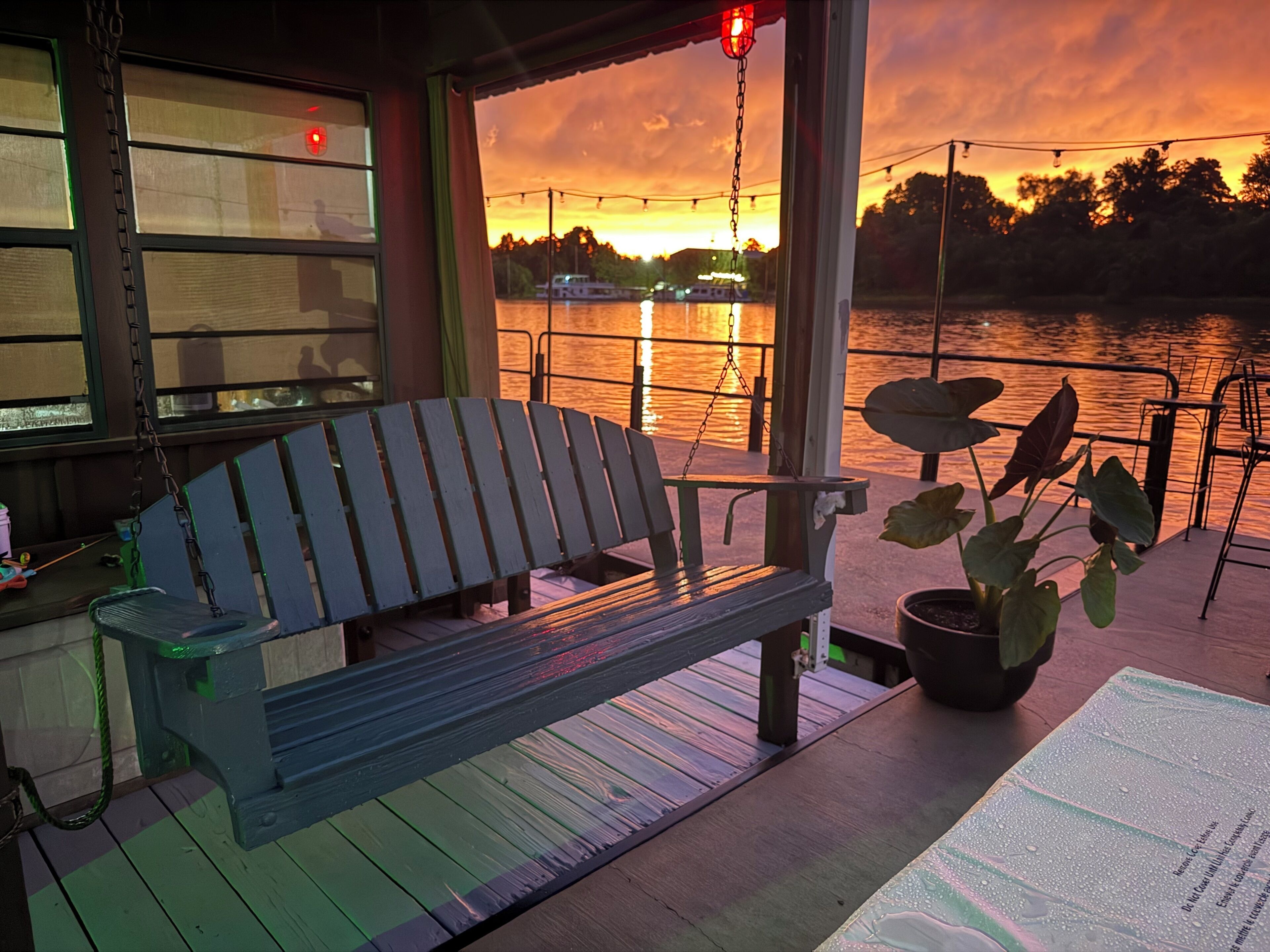 Une terrasse en bois avec un banc et un pot de fleurs, donnant sur une étendue d’eau avec un coucher de soleil en arrière-plan.