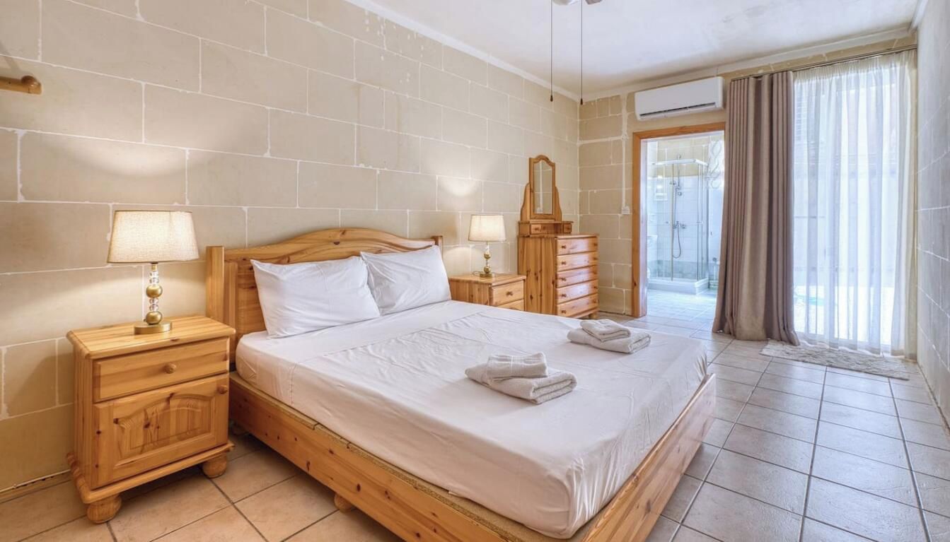 A hotel room with a wooden bed, a nightstand with a lamp, a dresser, and a view of the outside through a glass door.