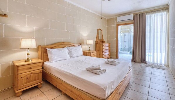 A hotel room with a wooden bed, a nightstand with a lamp, a dresser, and a view of the outside through a glass door. - Stone Apt in Nadur | Terrace & Patio | Sleeps 6 (Nadur)
