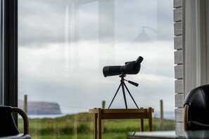 Een telescoop op een statief voor een raam, met uitzicht op een bewolkte lucht en een veraf gelegen landschap.