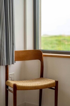 Une chaise en bois, dotée d’un siège en osier, est placée près d’une fenêtre qui offre une vue sur un paysage verdoyant.