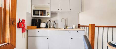 Cocina compacta con armarios blancos, microondas, hervidor de agua y cafetera.