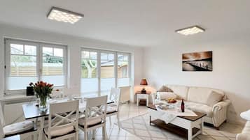 A spacious living area with a dining table, white chairs, a sofa, and a coffee table. There are large windows providing natural light and a view of the outside.