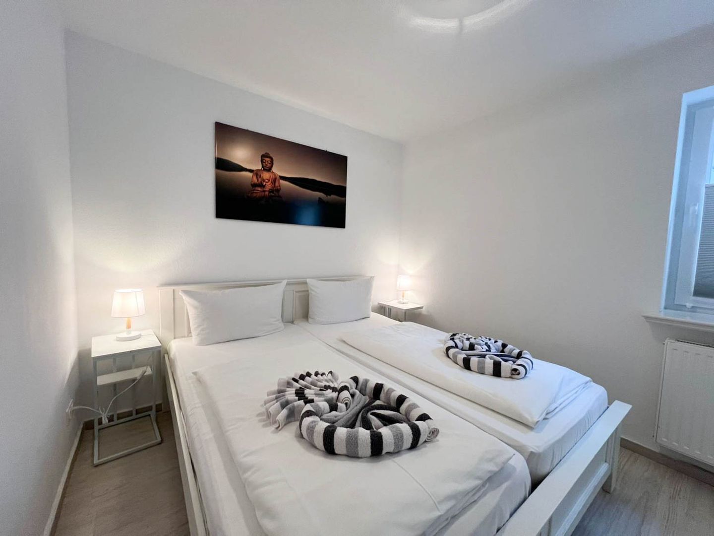 A hotel room with two beds, a nightstand with a lamp, a wall-mounted picture, and a window.