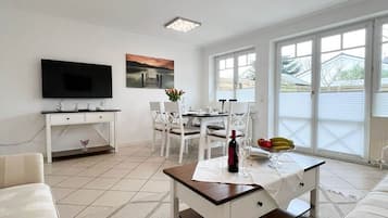 A modern living room with a flat-screen TV, a dining area with a table and chairs, and a large window with blinds.