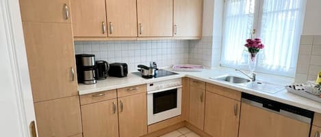 A compact kitchen with light wooden cabinets, a white countertop, a built-in oven, and a window with sheer curtains.