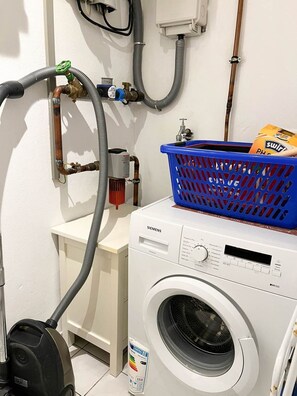 Ein Waschraum mit einer weißen Siemens Waschmaschine, einem blauen Wäschekorb und einem Staubsauger.