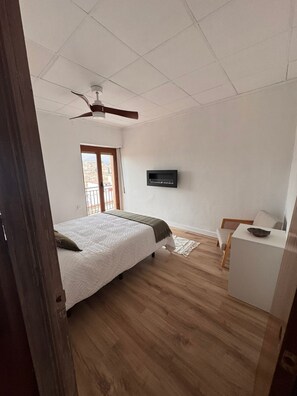 A bedroom with a bed, a ceiling fan, a TV mounted on the wall, and a wooden floor.