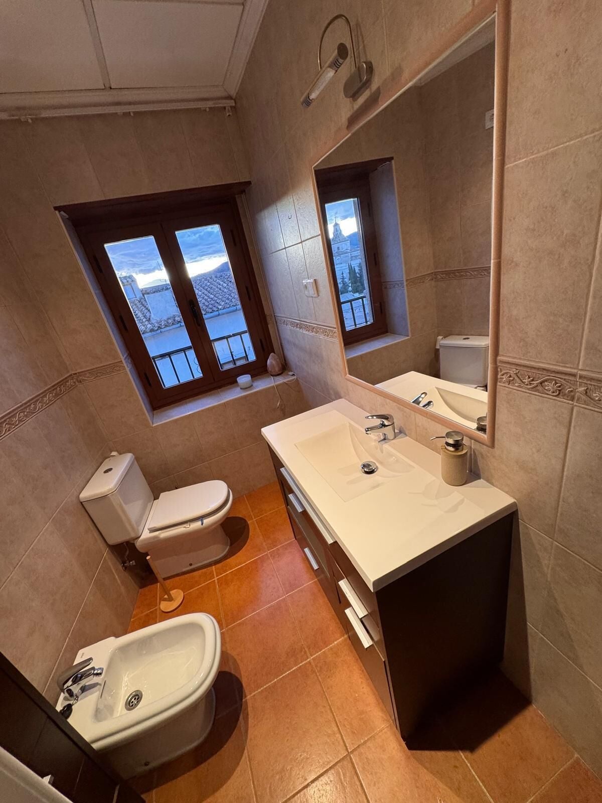 Un baño con inodoro, bidé, lavamanos y una ventana con vista a un edificio.