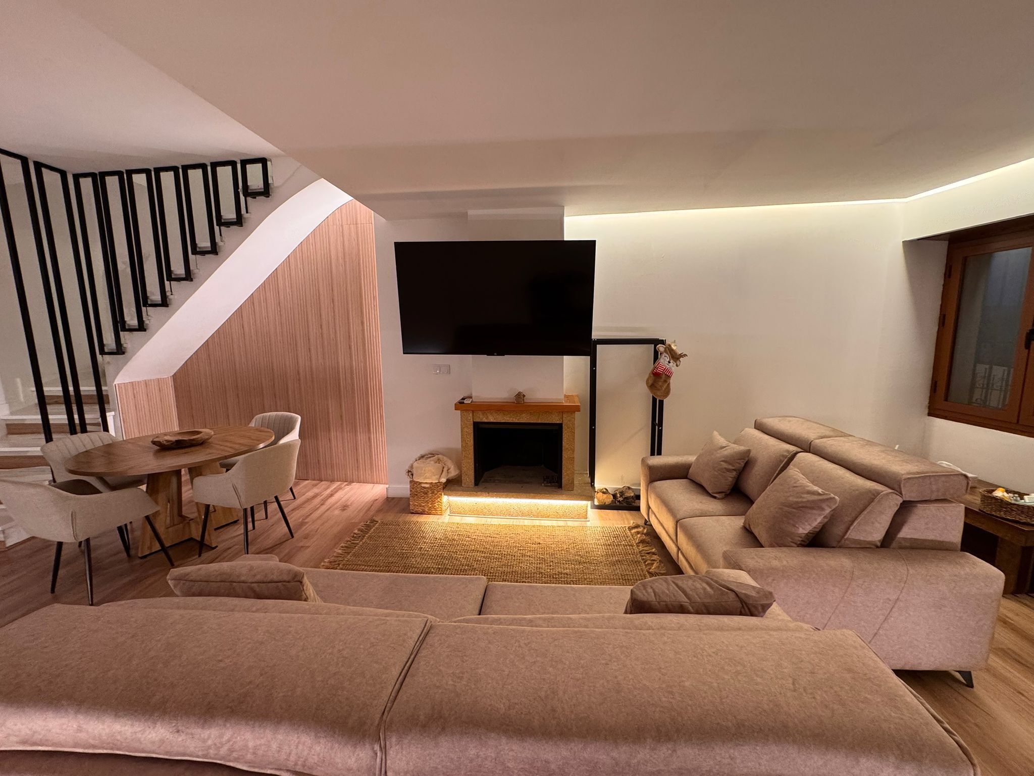 A modern living room with a sofa set, a dining table with chairs, a flat-screen TV mounted on the wall, and a staircase with black railings.