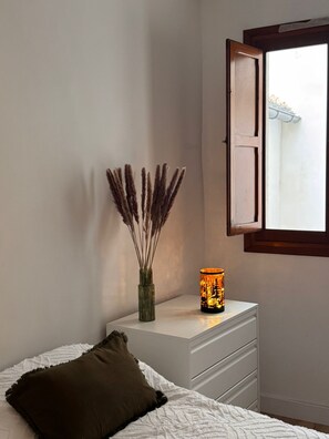 A bedroom with a bed, a bedside table with a lamp, a window, and a wall-mounted shelf with dried plants.