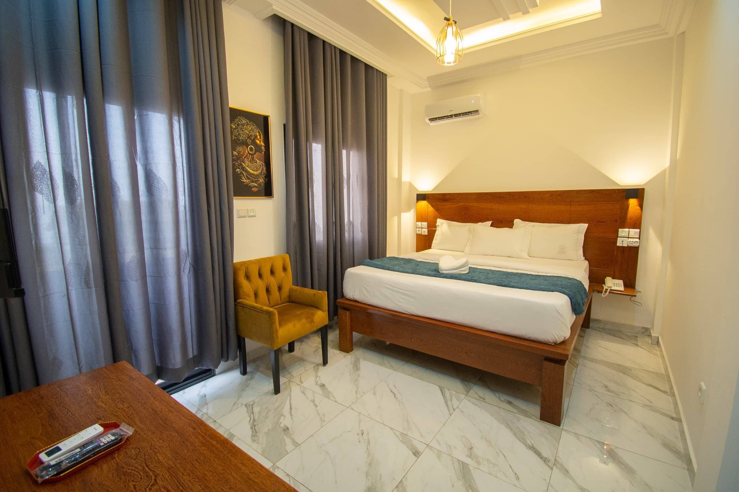 Quarto de hotel com cama, uma cadeira, escrivaninha e uma janela grande com cortinas.