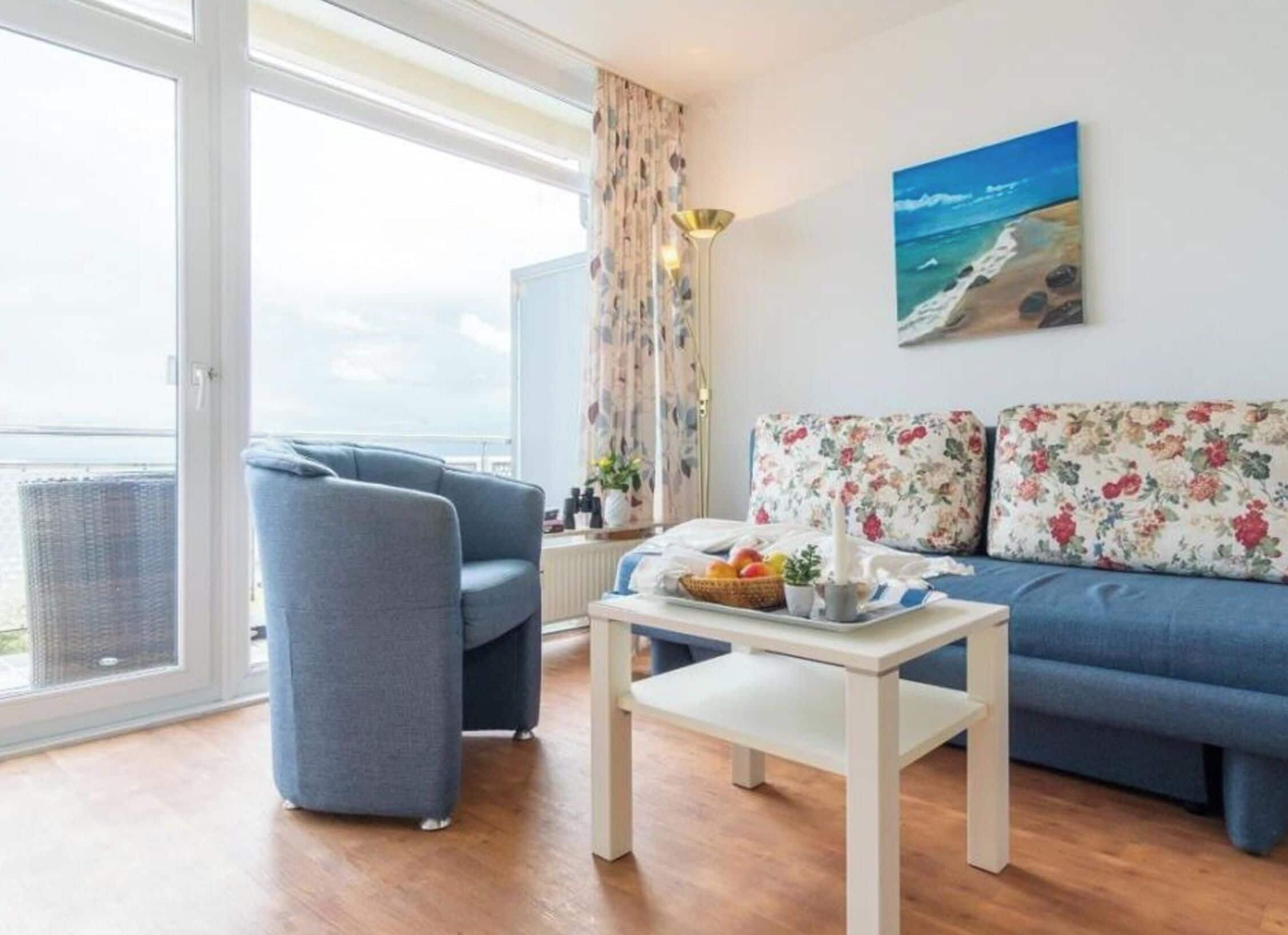Una sala de estar con un sofá, una silla, una mesa con una bandeja de frutas y un cuadro de una escena de playa.