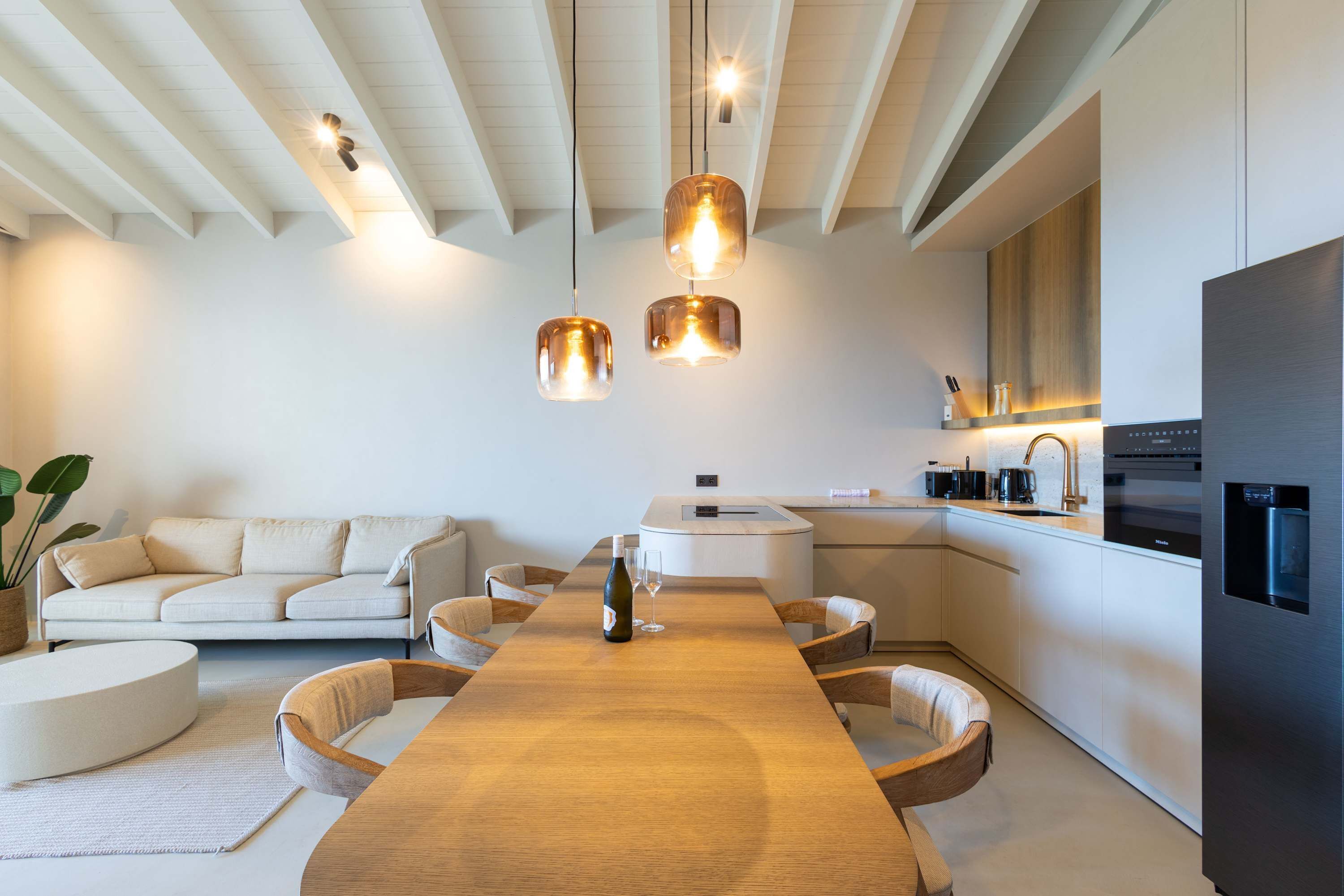 Eine moderne Küche mit einer zentralen Kochinsel, einem Essbereich mit Holztisch sowie einer Sitzecke inklusive Sofa und Sesseln.