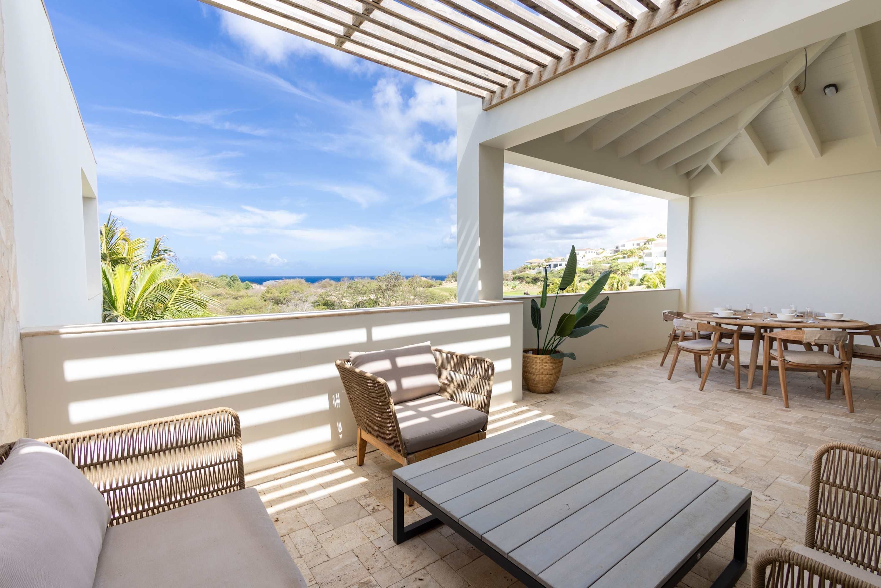 Une terrasse extérieure ensoleillée, avec un canapé, une table et des chaises, offrant une vue sur un paysage côtier.