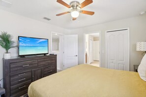 Une chambre à coucher avec un lit, un ventilateur de plafond, un téléviseur posé sur une commode et une porte donnant sur une autre pièce.
