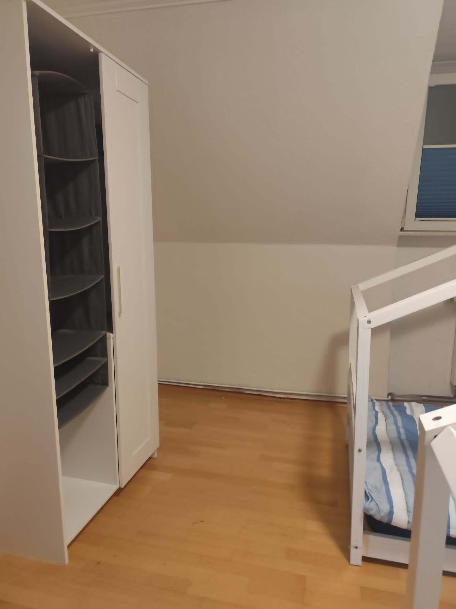 A room with a white wardrobe, a wooden floor, and a white table with a striped cushion.