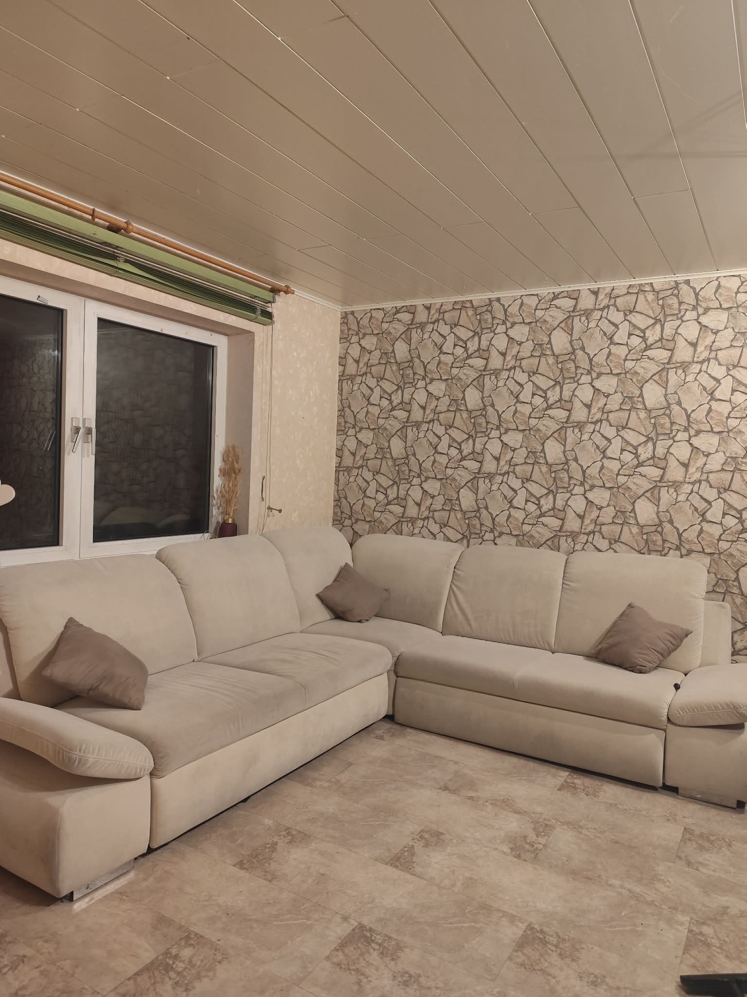 Ein beiges Ecksofa, eine Steinwand und ein Fenster mit Jalousien.
