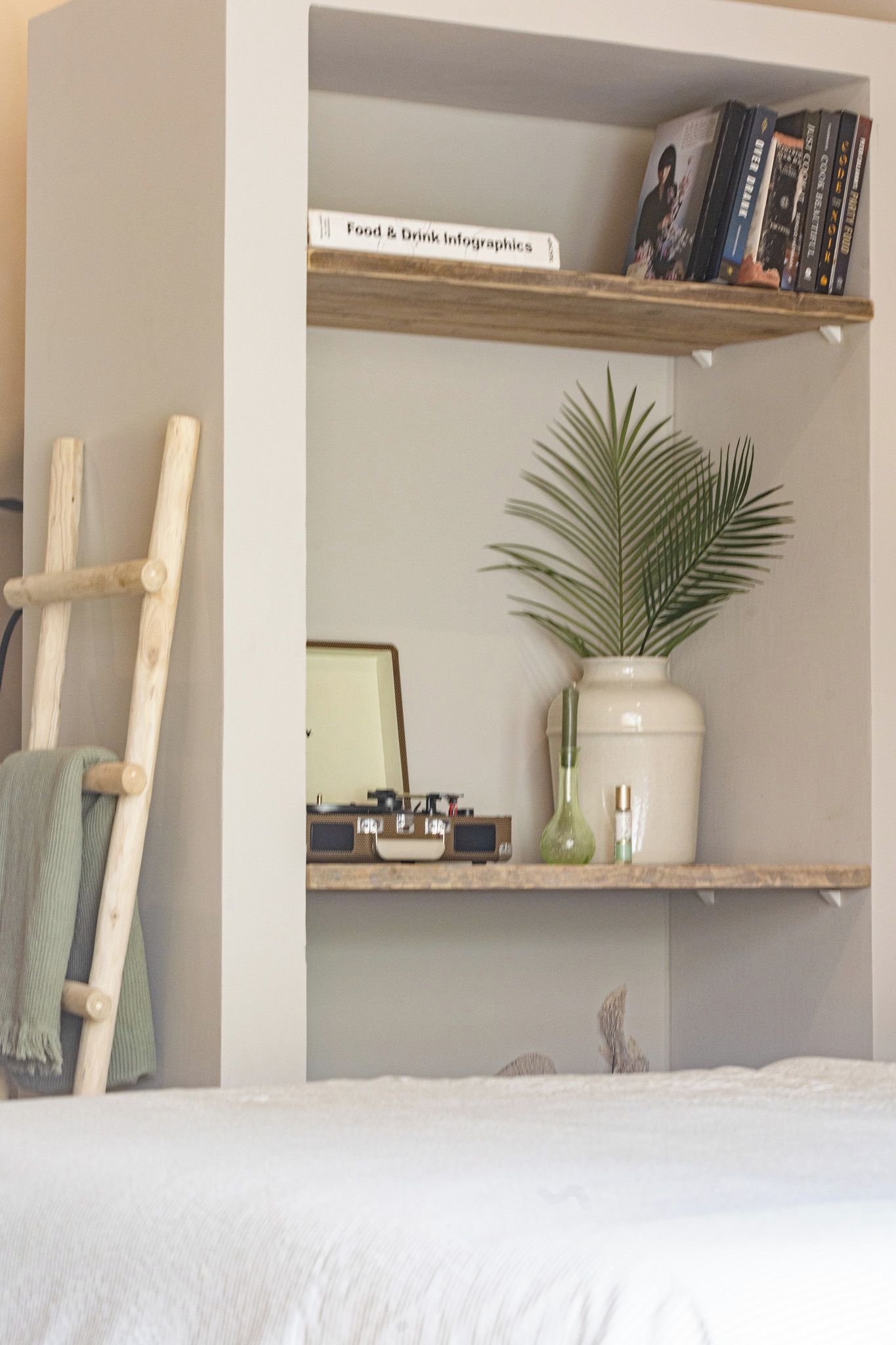 A bedroom with a bed, a wooden ladder, a shelf with books, a plant, and a framed picture.