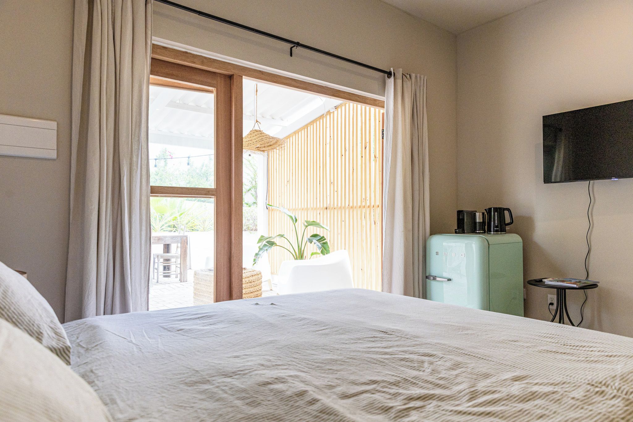 Habitación de hotel con cama, televisor montado en la pared, kitchenette con refrigerador y puerta corrediza de vidrio que da a un área exterior.