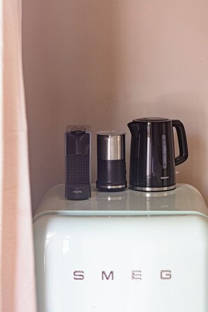 Ein Küchenbereich mit einem weißen Gerät mit der Aufschrift 'SMEG' und drei Gegenstände darauf: einem schwarzen Wasserkocher, einer schwarzen Kaffeemaschine und einem schwarzen elektrischen Milchaufschäumer.
