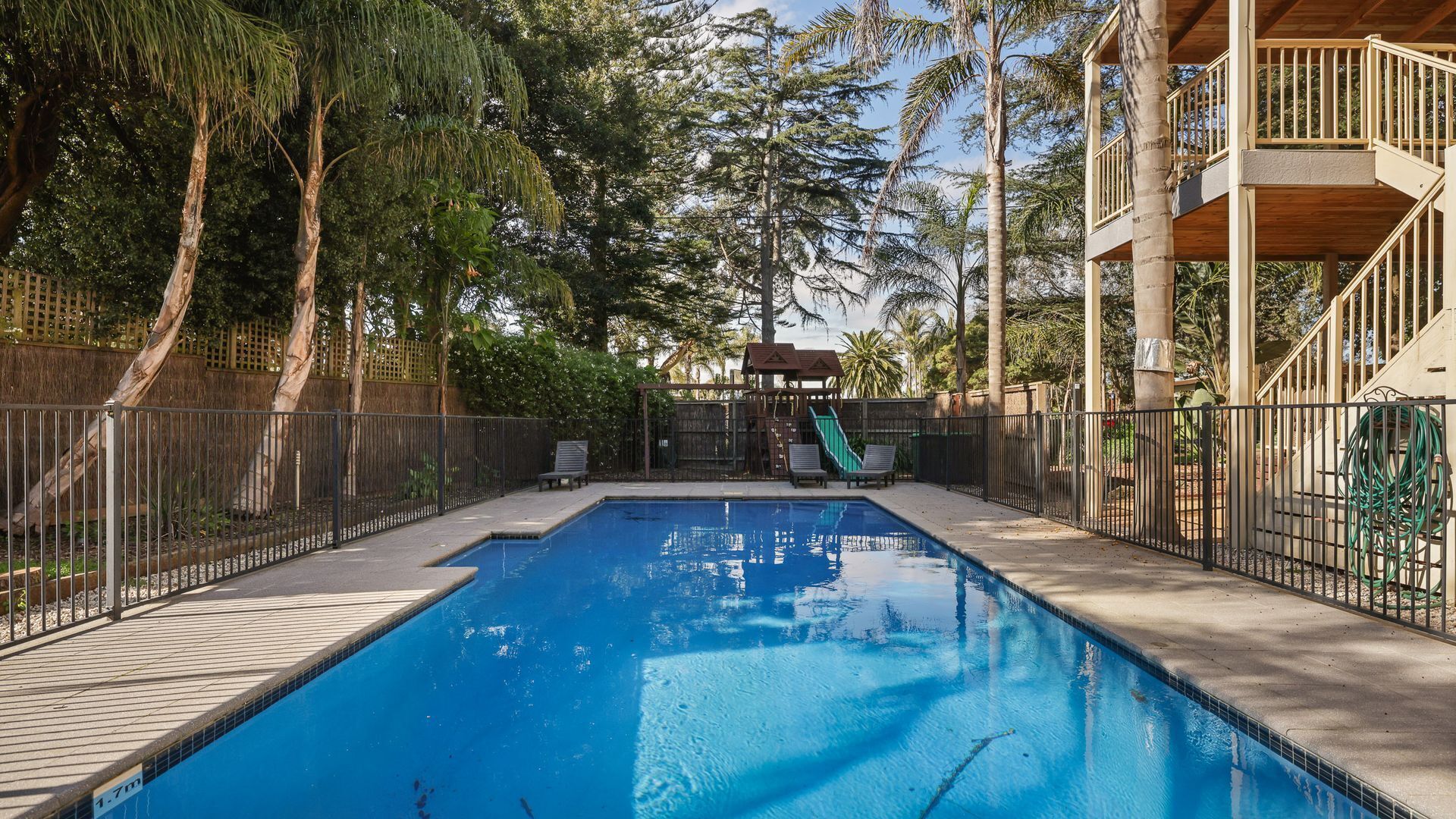 Une piscine avec une terrasse en bois, entourée d’une clôture, et une structure de jeux pour enfants en arrière-plan.