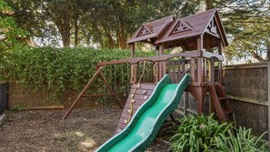A wooden play structure with a slide and climbing area, surrounded by a fence and greenery. - Unit 1 PI Holiday Retreat (Cowes)