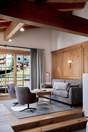 Ein Wohnzimmer mit Sofa, Sessel und Couchtisch, Holzbalken an der Decke und Blick durch eine Glastür.