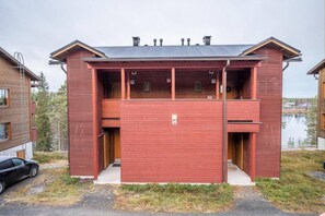 A red building with two balconies, a car parked in front, and a body of water in the background. - Länsirinne C9 | Modern 2BR Apt with Lake Views (Ruka)