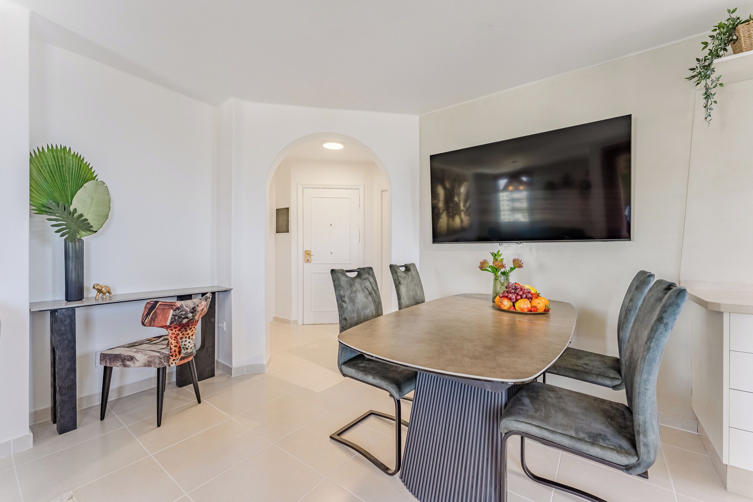 Een moderne eetruimte met een ronde tafel, gestoffeerde grijze stoelen, een vaas met bloemen en een flatscreen televisie aan de muur.