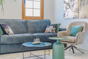 Ein Wohnzimmer mit einem grauen Sofa, einem beigefarbenen Sessel, einem kleinen runden Tisch mit Glasplatte und einer grünen Vase.