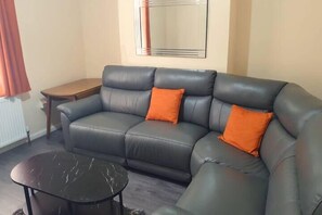 A living room with a sofa, two orange cushions, a small round table, and a mirror on the wall. - 5 Bed House Manchester - Sleeps 9 - Free Parking (Manchester)