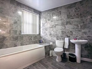 A bathroom with a bathtub, toilet, and sink, all with white fixtures, set against a grey tiled wall. - 5 Bed House Manchester - Sleeps 9 - Free Parking (Manchester)