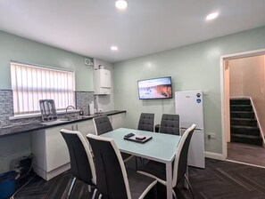 A compact kitchen with a dining area, a TV mounted on the wall, and a refrigerator. - 5 Bed House Manchester - Sleeps 9 - Free Parking (Manchester)