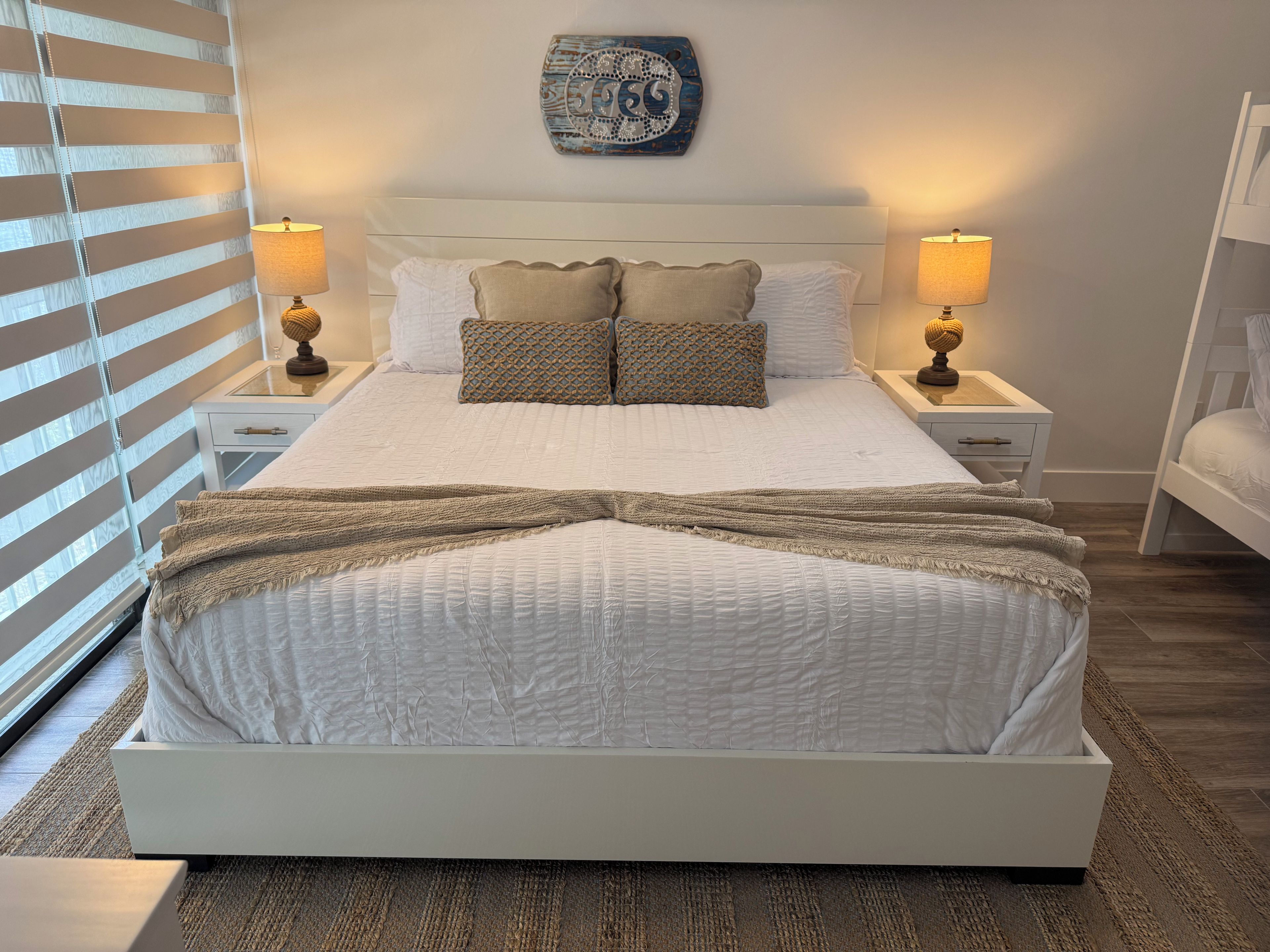 Cama bem arrumada com cabeceira branca, duas mesas de cabeceira com abajures, um objeto decorativo na parede e uma janela com persianas.